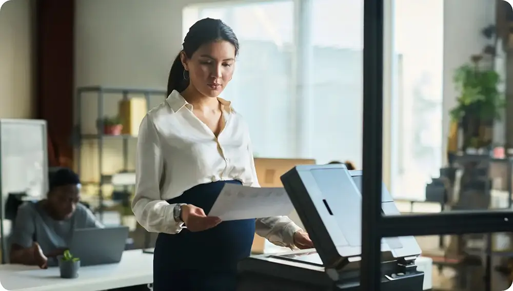 Businesswoman scanning documents at a copier in a busy office environment.