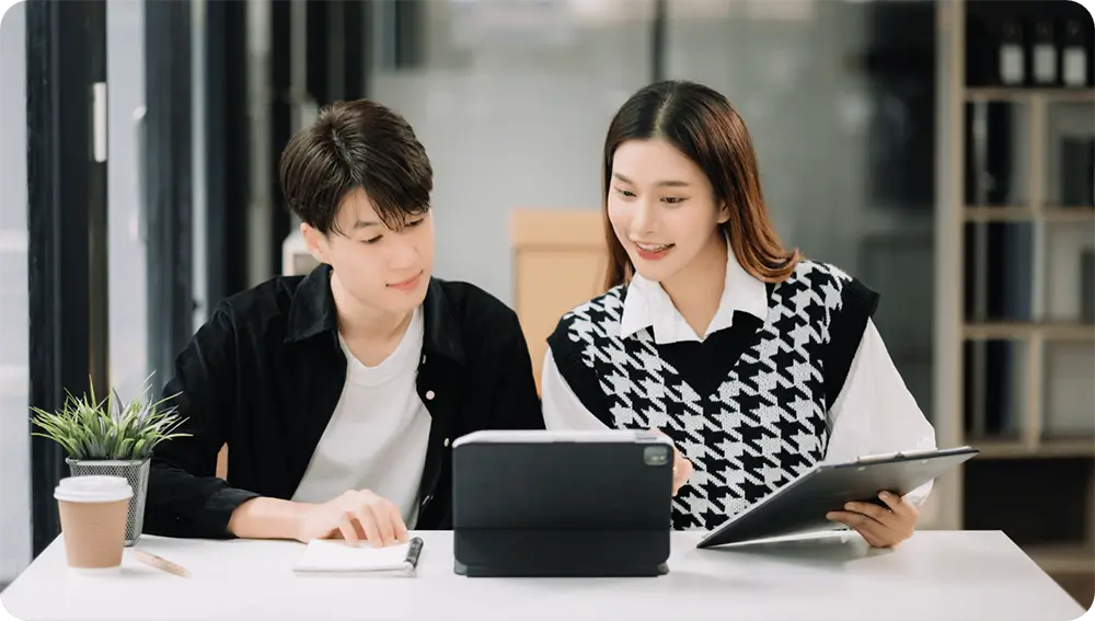 Two young professionals collaborate at a desk, one taking notes while the other shows something on a tablet, both smiling.